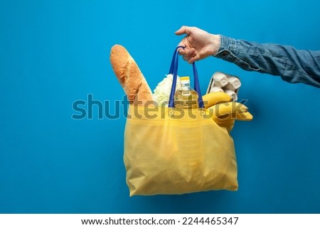 fabric yellow eco bag full of food holds a guy's hand on a blue background, food delivery concept, plastic rejection concept