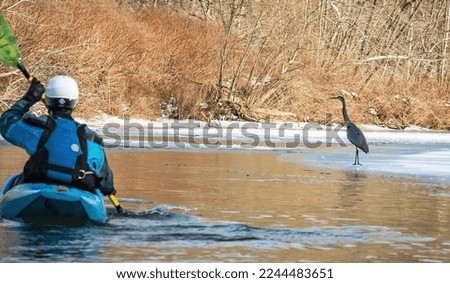 Newark, Ohio, USA - 12-28-2022 - Kayaker Paddling in Winter with ice on river and snow, with Great Blue Heron Standing on ice.  Royalty-Free Stock Photo #2244483651