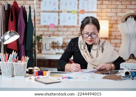 Asian middle-aged female fashion designer working in a studio by idea drawing sketches with colorful thread and sewing for a dress design collection, professional boutique tailor SME entrepreneur.