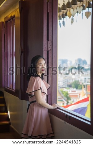 Portrait picture an Asian woman in a pink dress, she is looking at a camera  standing next to a window in temple while traveling in Asia.