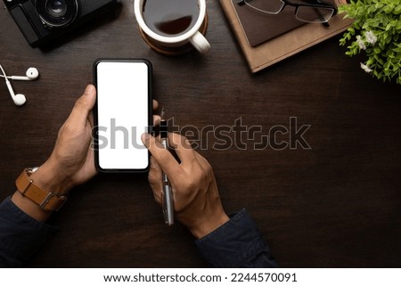 Above view of businessman hand holding pen and using smart phone on wooden table.