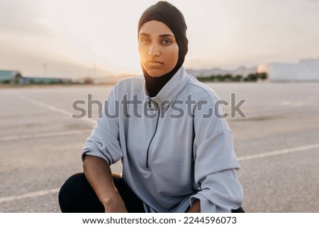 Sportswoman with a hijab looking at the camera while squatting outdoors. Athletic Muslim woman taking a break during her morning workout session. Royalty-Free Stock Photo #2244596073