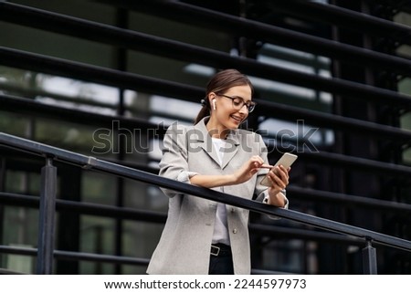 A young businesswoman elegantly dressed leaning on the railing outdoors and listening to music or podcast over her phone. A businesswoman on a break