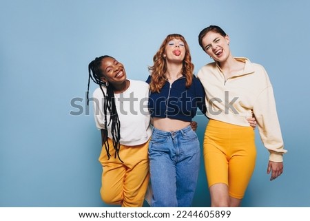 Cheerful female friends making funny faces and looking at the camera while embracing each other. Group of happy young women having fun while standing against a blue studio background. Royalty-Free Stock Photo #2244605899