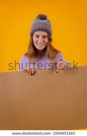 A woman in a wool cap holding and pointing at a cardboard sign on a yellow background