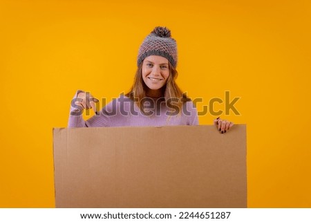 Portrait of a woman in a wool cap pointing at a cardboard sign on a yellow background