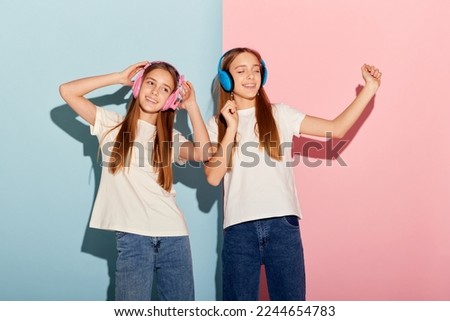 Music. Delighted young girls, two sisters friends in colorful headphones isolated over pink-blue studio background. Concept of modern youth fashion, relationship, emotions, beauty and youth.