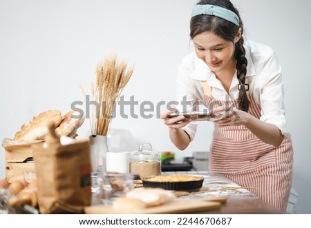 Young woman wears apron taking selfie photo with homemade pie in kitchen. Portrait of beautiful Asian female baking dessert and having fun taking photo by smartphone for online social. Home cookery. Royalty-Free Stock Photo #2244670687