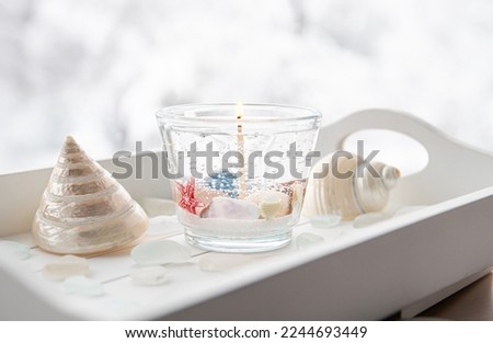 Homemade underwater seashell clear gel wax candle inside clear cup. Beautiful candle on white wood tray with sea glass and seashells on window sill at home. Royalty-Free Stock Photo #2244693449