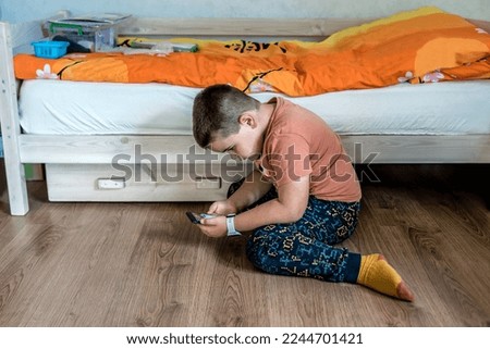 Little boy web surfing online on smartphone, watching videos or playing games, sitting on wooden laminate floor at home