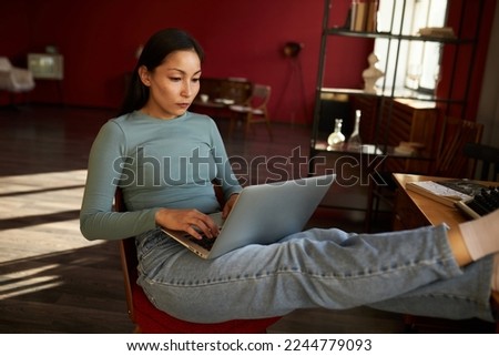 A concentrated girl sits on a chair with a computer, her legs are on the table, typing a text or talking on the Internet through instant messengers on work issues Royalty-Free Stock Photo #2244779093