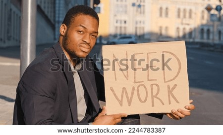 Fired sad unemployed businessman ethnic guy in business suit in city looking for job. Millennial upset African American man male searching employment in crisis sitting outdoors with poster Need Work