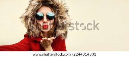 Portrait of stylish woman blowing her lips sends air kiss stretching hand for taking selfie with smartphone wearing jacket with fur hood