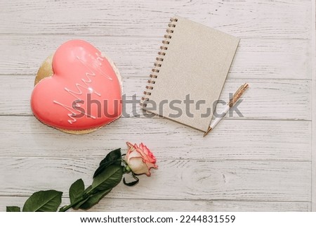 Heart shaped pink mousse cake on table with rose and notepad.