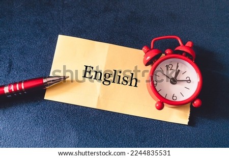 The word English on a yellow piece of paper with a red alarm clock and pen in the composition.
