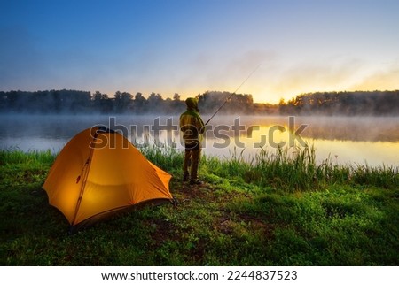 Fisherman on the bank of a foggy river near an orange tent in the early morning Royalty-Free Stock Photo #2244837523
