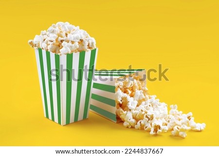 Two green white striped carton buckets with tasty cheese popcorn, isolated on yellow background. Box with scattering of popcorn grains. Movies, cinema and entertainment concept.