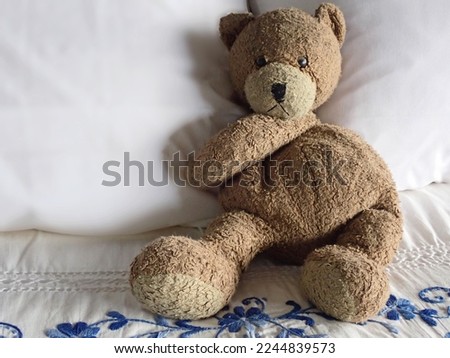 Shadow and light play on shaggy beloved brown stuffed teddy bear with round belly and arms crossed under tan pouting snout and shiny black eyes on crisp white bed pillows with blue embroidery sheet.