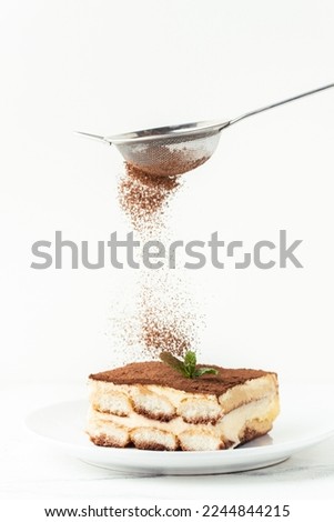 Pastry Chef sprinkles cocoa powder through a sieve on a tiramisu cake decorated with green mint. Making traditional Italian no-baking dessert. White background Royalty-Free Stock Photo #2244844215