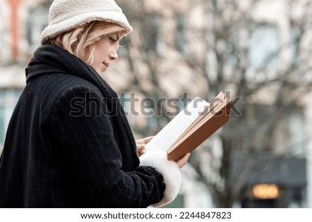 Portrait of young woman reading a book in an urban environment. Cloudy winter day and she is outdoors wearing black coat, scarf and whit cap. Royalty-Free Stock Photo #2244847823