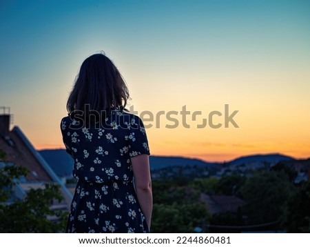 Young black hair girl in a floral dress looking at a colorful city sunset from the back