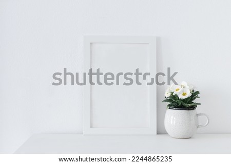 Spring, Easter interior still life composition. Blank picture frame mockup on white table. White, yellow potted primroses in coffee cup flower pot. White wall background. Minimal Scandinavian interior