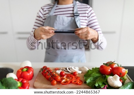 Female hands holding modern cell phone, cropped of woman in apron taking photo of various fresh organic vegetables on table while preparing healthy meal at home, kitchen interior. Food blogging