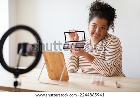 Cute attractive mixed race young woman plus size beauty blogger showing followers brand new eye shadow palettes, lady influencer streaming via cell phone while unpacking and testing cosmetics at home