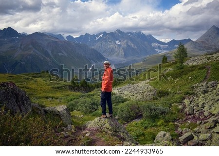Hiker wearing an orange anorak, blue trousers and khaki cowboy hat tursn his head back while making a photo with splendid mountain background. Man on a hiking walk in the Altai Mountains of Kazakhstan Royalty-Free Stock Photo #2244933965