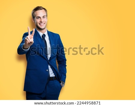 Excited businessman in confident suit showing two fingers or victory sign gesture, on vivid yellow colour background. Handsome happy man. Copy space for ad, slogan or text.