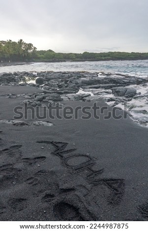 A vertical photo of a black sand beach at Punaluʻu Beach on Big Island of Hawaii. With the words ALOHA spelled out in the sand and waves hitting the lava rocks