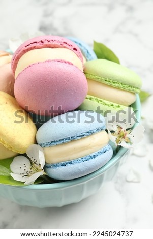 Delicious colorful macarons and flowers in bowl on white table, closeup
