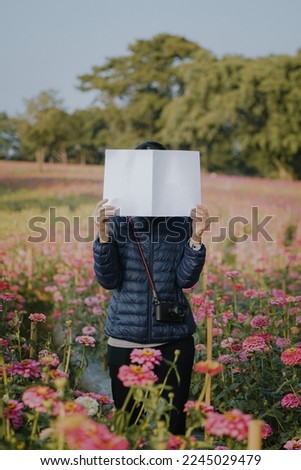 Teenage girl covering her face with blank book or magazine with zinnia flowers field.