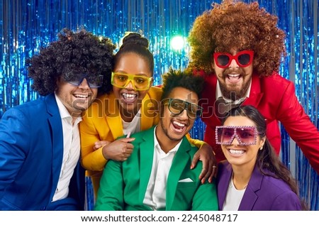 Happy cheerful joyful people wearing different bright yellow green, red, purple costumes, party glasses and funny crazy wigs smile and look at camera on glittering blue foil fringe photobooth backdrop Royalty-Free Stock Photo #2245048717