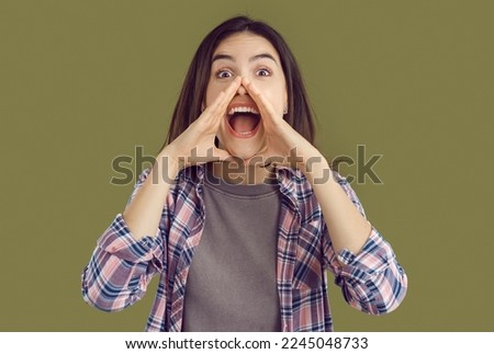 Shouting excited young brunette woman in plaid shirt with hands near mouth on khaki background. Positive emotions concept. Banner with shouting woman calling somebody or distributing information. Royalty-Free Stock Photo #2245048733