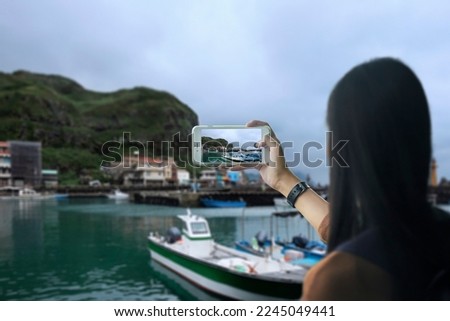 Back view portrait of a woman taking photo of fishing village full of anchored fishing boat in cloudy day by her smart phone 