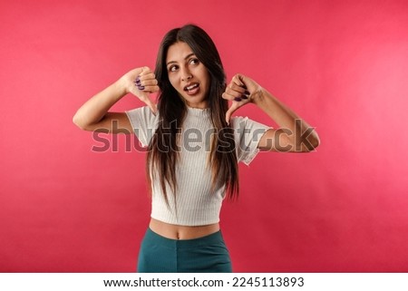 Brown-haired woman wearing white ribbed crop isolated over red background makes a thumbs down gesture with both hands. Not satisfied and looks at the camera.