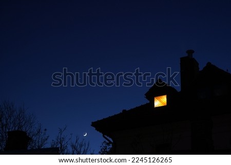 Silhouette of house roof with chimney and lit attic small window at night against clear sky with moon and star Royalty-Free Stock Photo #2245126265