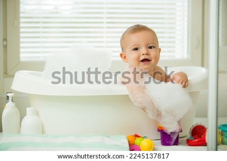 Cute little baby sitting in white bathtub with foam and soap bubbles. Taking bath and playing with toys. Baby hygiene.  Royalty-Free Stock Photo #2245137817
