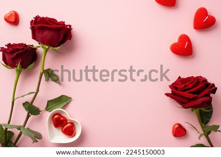 Valentine's Day concept. Top view photo of red roses heart shaped candles and saucer with chocolate candies on isolated pastel pink background with copyspace