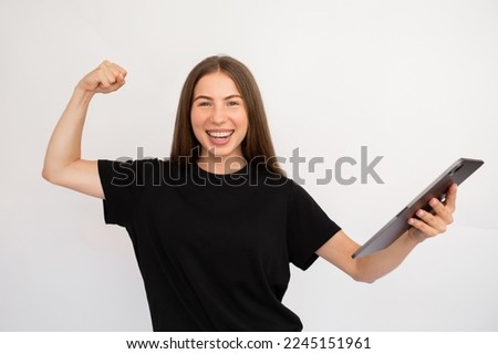 Portrait of happy young woman with touchpad celebrating success. Caucasian lady wearing black T-shirt making winning gesture and smiling at camera. Wireless technology and success concept