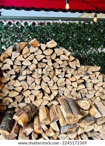 Wood logs stacked one on top of each other in front of white fence covered in artificial plant leaf and hanging outdoor string lights.