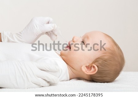 Doctor hand in white rubber protective gloves holding and giving plastic tube of rotavirus vaccine. Baby boy lying down on changing table and receiving medicine. Side view. Closeup. Royalty-Free Stock Photo #2245178395