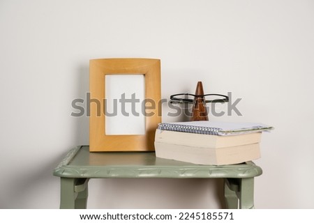 empty picture frame mock up with soft backed books wooden glasses stand on green table plain background 