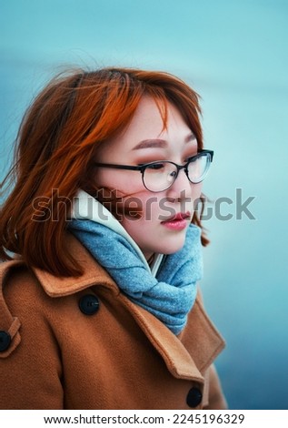 Portrait of a beautiful Asian woman with glasses, wearing a blue scarf and a beige coat against the background of a cold sea on a winter day. The romance of travel. The cold season.