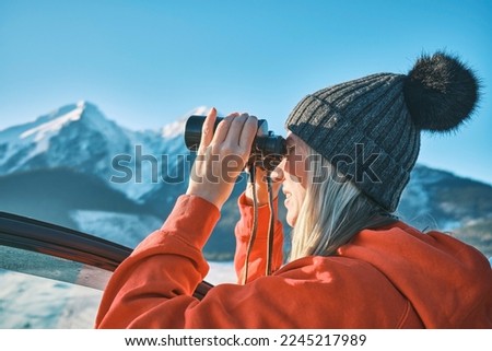 Woman traveling exploring, enjoying the view of the mountains, landscape, lifestyle concept winter vacation outdoors. Female standing near the car in sunny day, Girl looking through binoculars, travel