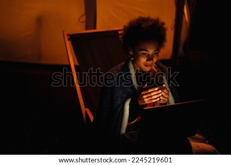 Young african woman with blanket working on laptop and drinking hot tea while sitting on chair near glamping tent in nature
