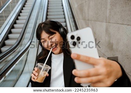Young asian woman taking selfie photo on cellphone and holding coffee while riding escalator