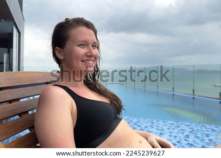 Happy positive beautiful girl, young woman is enjoying summer vacation in swimming pool, luxury spa on roof of hotel in exotic tropical country with sea, ocean view, breath deep deeply fresh air.