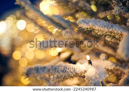 Christmas tree covered snow with yellow garlands lights and bokeh copy space, outdoor xmas green tree with decorative garlands, outdoor winter holiday atmosphere. Festive Christmas tree decorations